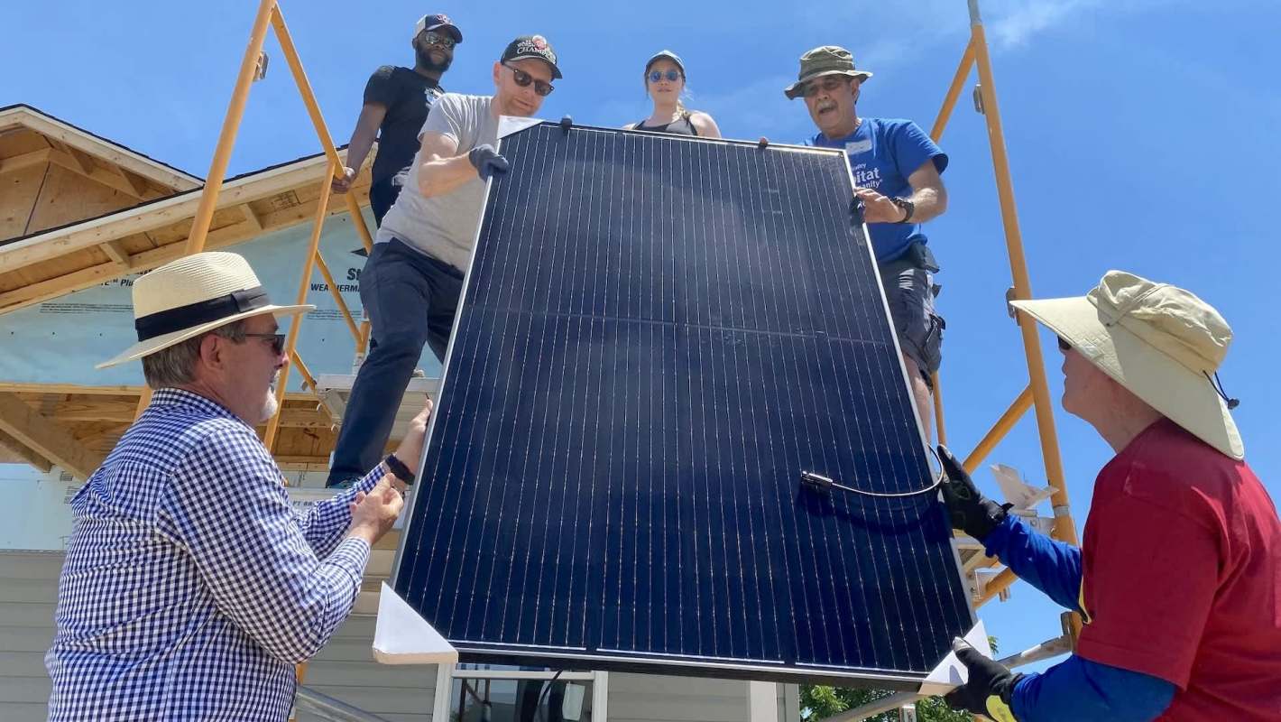 A group of people working together to install a solar panel on a building under a clear blue sky.