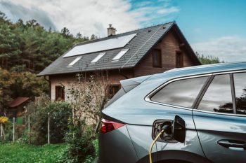 An electric vehicle is charging beside a modern house with solar panels on the roof, surrounded by greenery.
