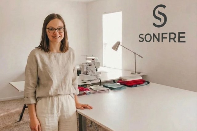 A woman in a light beige outfit stands in a workspace with sewing equipment and fabric stacks.