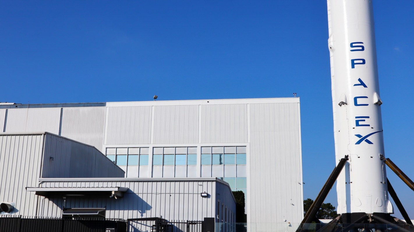 A close-up of a SpaceX rocket standing beside a large industrial building against a clear blue sky.