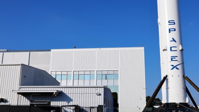 A close-up of a SpaceX rocket standing beside a large industrial building against a clear blue sky.