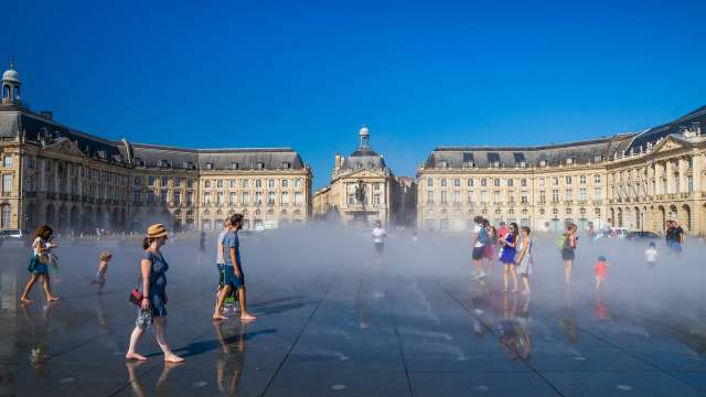 People play in a misty water installation in a square surrounded by historic architecture on a sunny day.