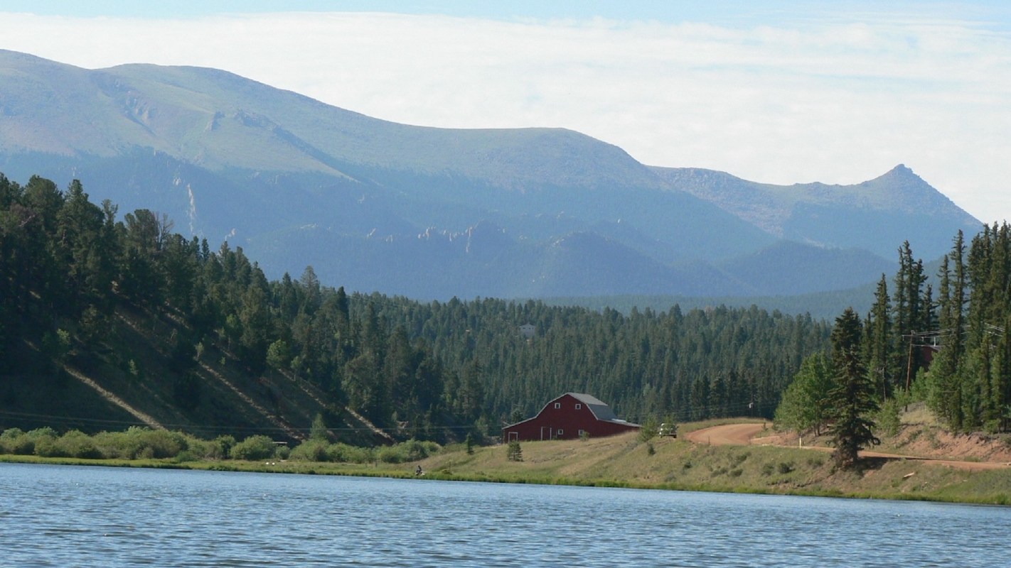 A red barn near a lake with mountains and forests in the background.