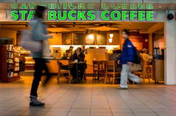 A busy Starbucks coffee shop with customers seated and people walking by in a modern interior setting.