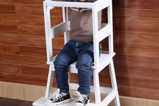 A young child sits in a white high chair against a wooden wall, wearing a sweater and jeans.