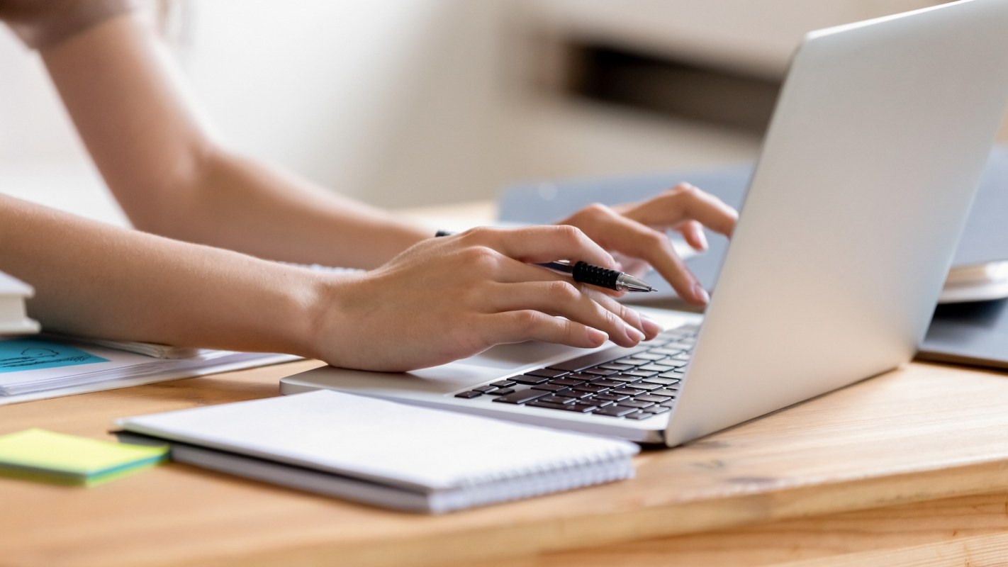 A person types on a laptop while holding a pen, with notebooks and sticky notes on a wooden desk.