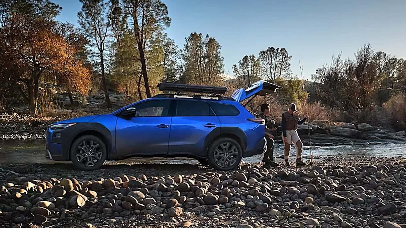 Two people with fishing gear next to an electric SUV parked on a rocky riverbank.