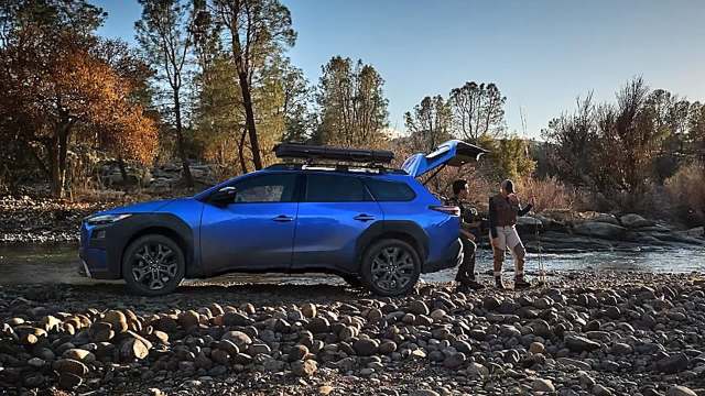 Two people with fishing gear next to an electric SUV parked on a rocky riverbank.