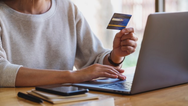A person holding a credit card while using a laptop on a wooden table with a notebook and smartphone nearby.