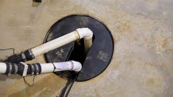 A view of a sump pump pit with two PVC pipes and a circular black lid on a concrete floor.