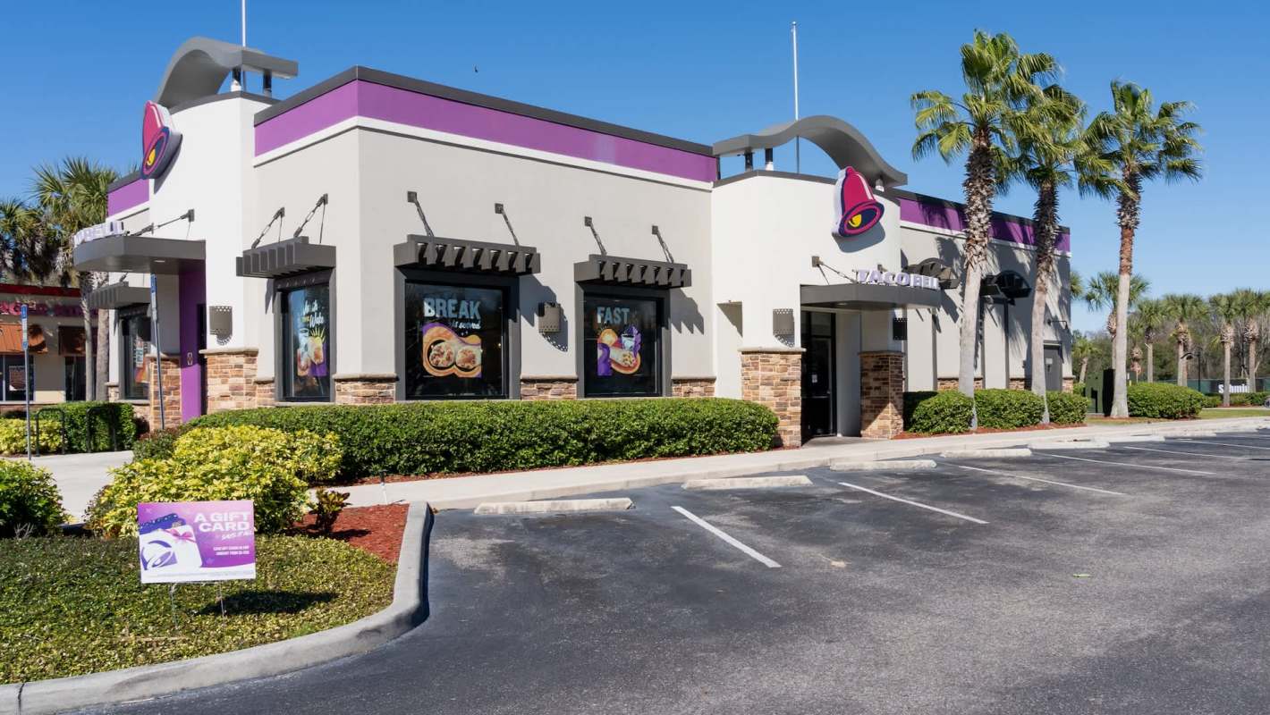 A Taco Bell restaurant with palm trees, clear blue sky, and promotional signage in front of an empty parking lot.