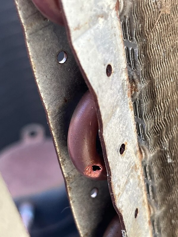 Close-up of a metal cooling coil with a hole, some rust and discoloration, revealing wear over time.