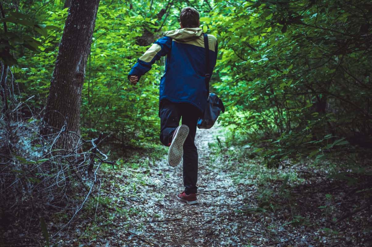 A person with a backpack jogs along a wooded trail surrounded by lush green foliage.