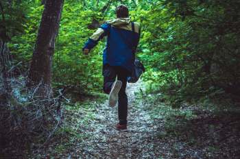 A person with a backpack jogs along a wooded trail surrounded by lush green foliage.