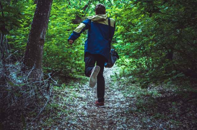 A person with a backpack jogs along a wooded trail surrounded by lush green foliage.