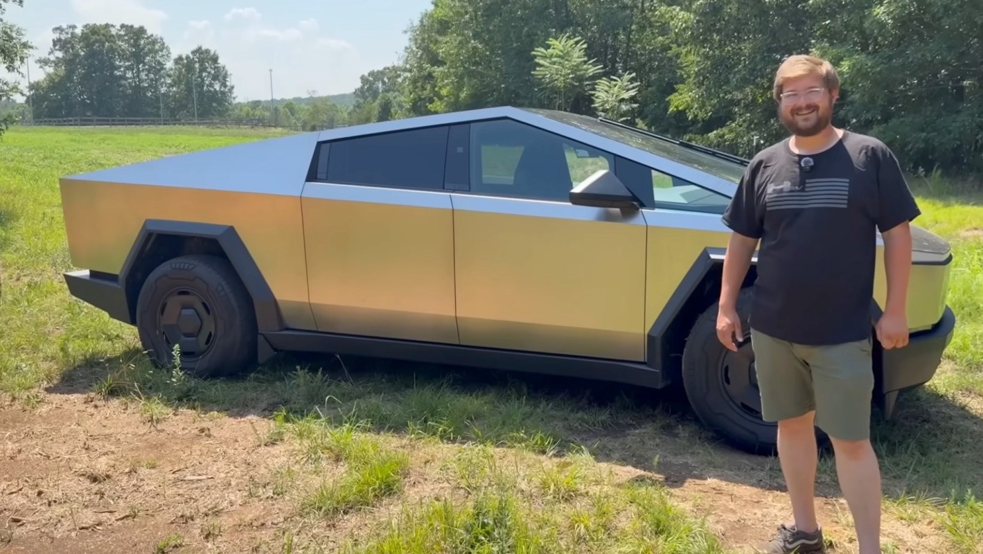 A man stands beside a Tesla Cybertruck in a grassy area with trees in the background.