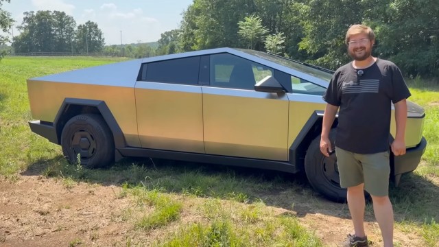 A man stands beside a Tesla Cybertruck in a grassy area with trees in the background.