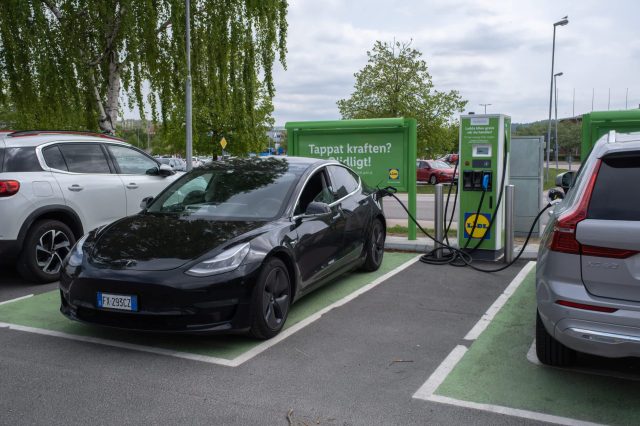 A black Tesla Model 3 charging at an electric vehicle station next to a silver SUV in a parking lot.