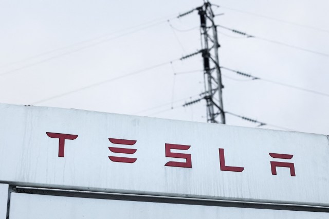 A close-up of a Tesla sign with a power line in the background under a cloudy sky.