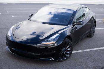 A black Tesla Model 3 parked in an empty lot, reflecting clouds on its sleek surface.