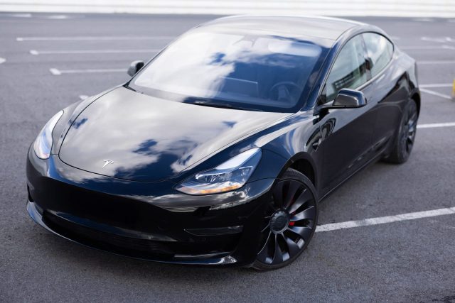 A black Tesla Model 3 parked in an empty lot, reflecting clouds on its sleek surface.