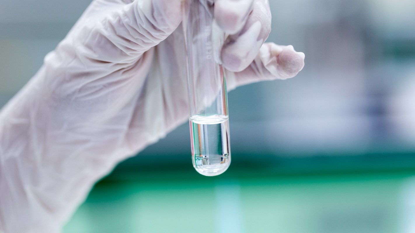 A gloved hand holds a test tube containing a clear liquid against a blurred laboratory background.