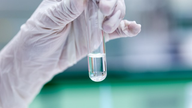 A gloved hand holds a test tube containing a clear liquid against a blurred laboratory background.