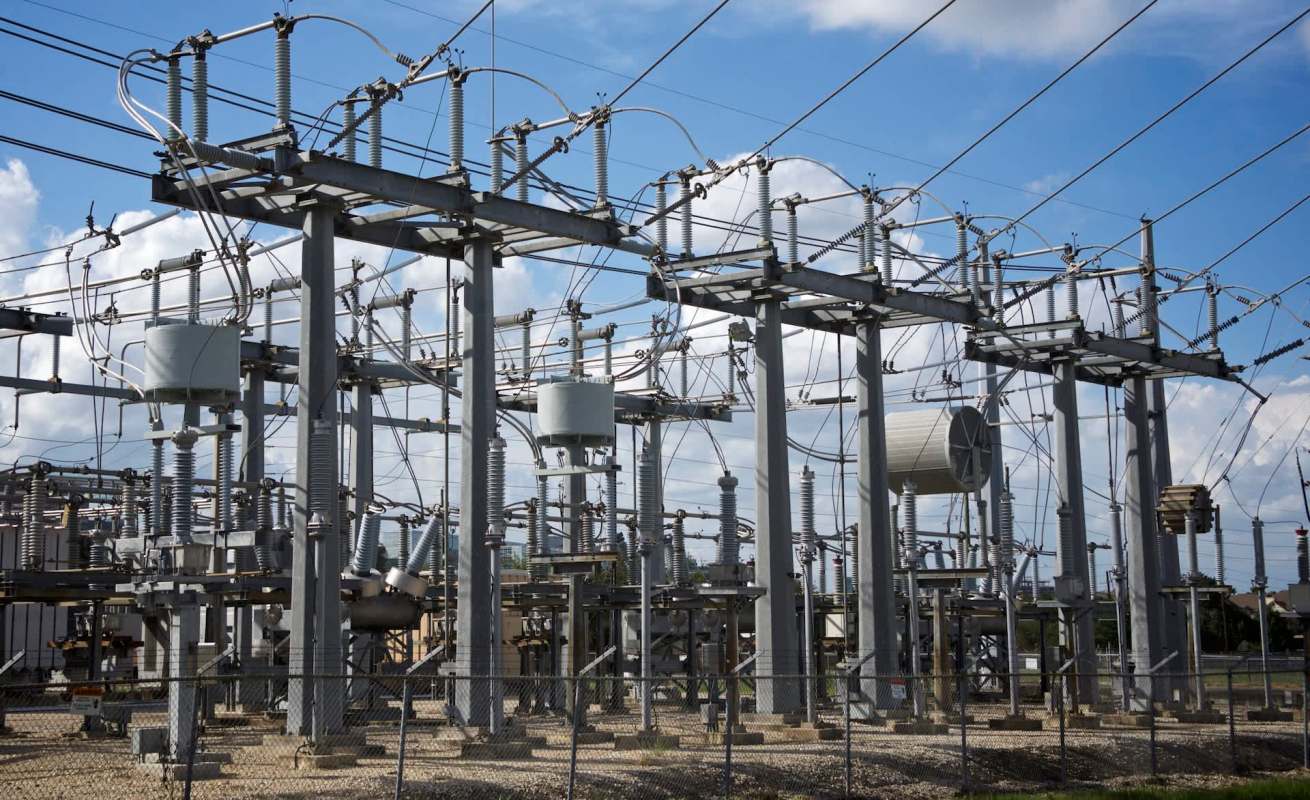 An electrical substation with metal structures, wires, and equipment under a cloudy sky.