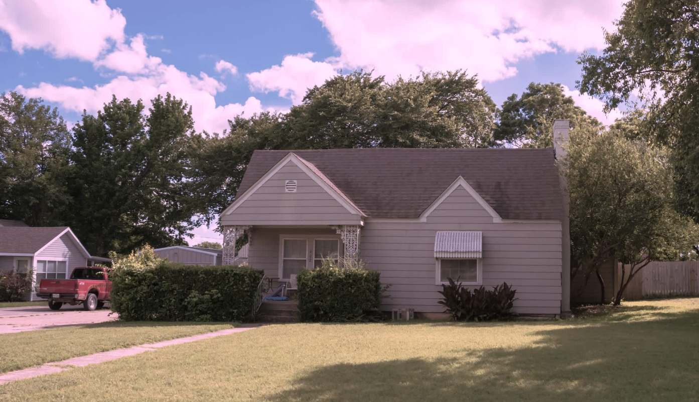 A small, light gray house with a covered porch, surrounded by greenery under a sunny blue sky.