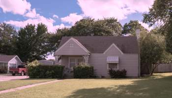 A small, light gray house with a covered porch, surrounded by greenery under a sunny blue sky.