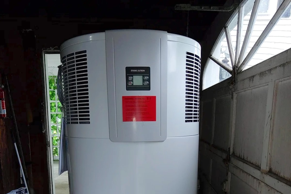 A large white heat pump water heater is positioned in a dimly lit garage next to a partially open door.