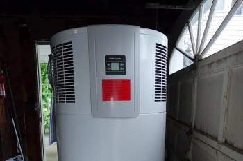 A large white heat pump water heater is positioned in a dimly lit garage next to a partially open door.