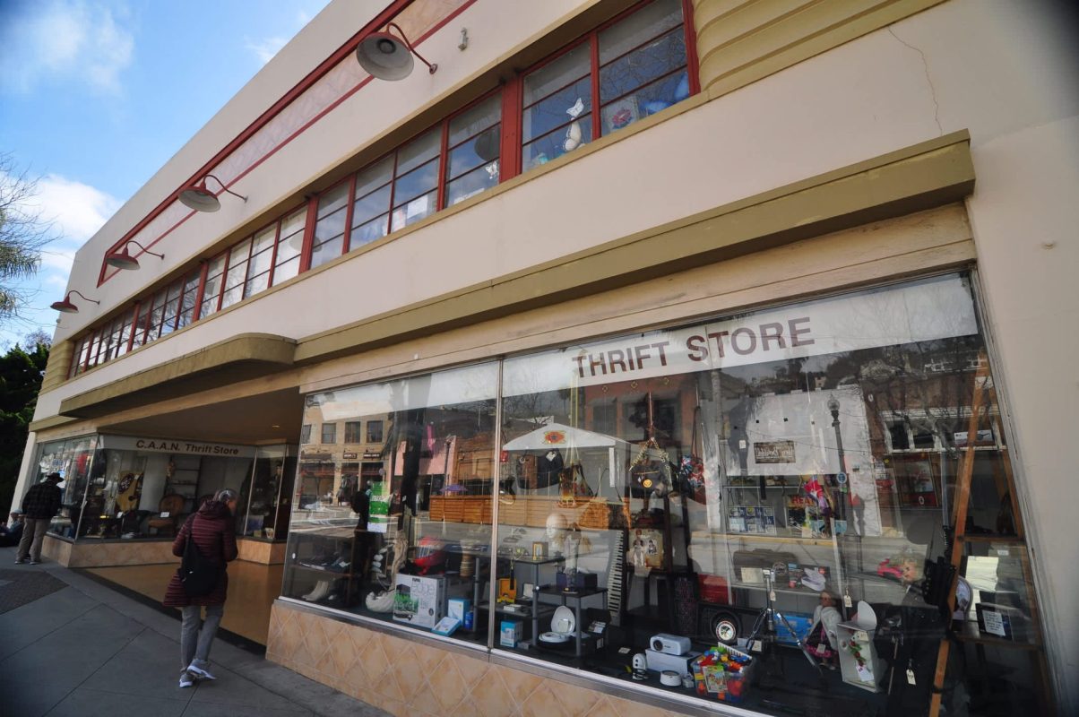 A thrift store with large windows displays various items, while shoppers walk by on the sidewalk.