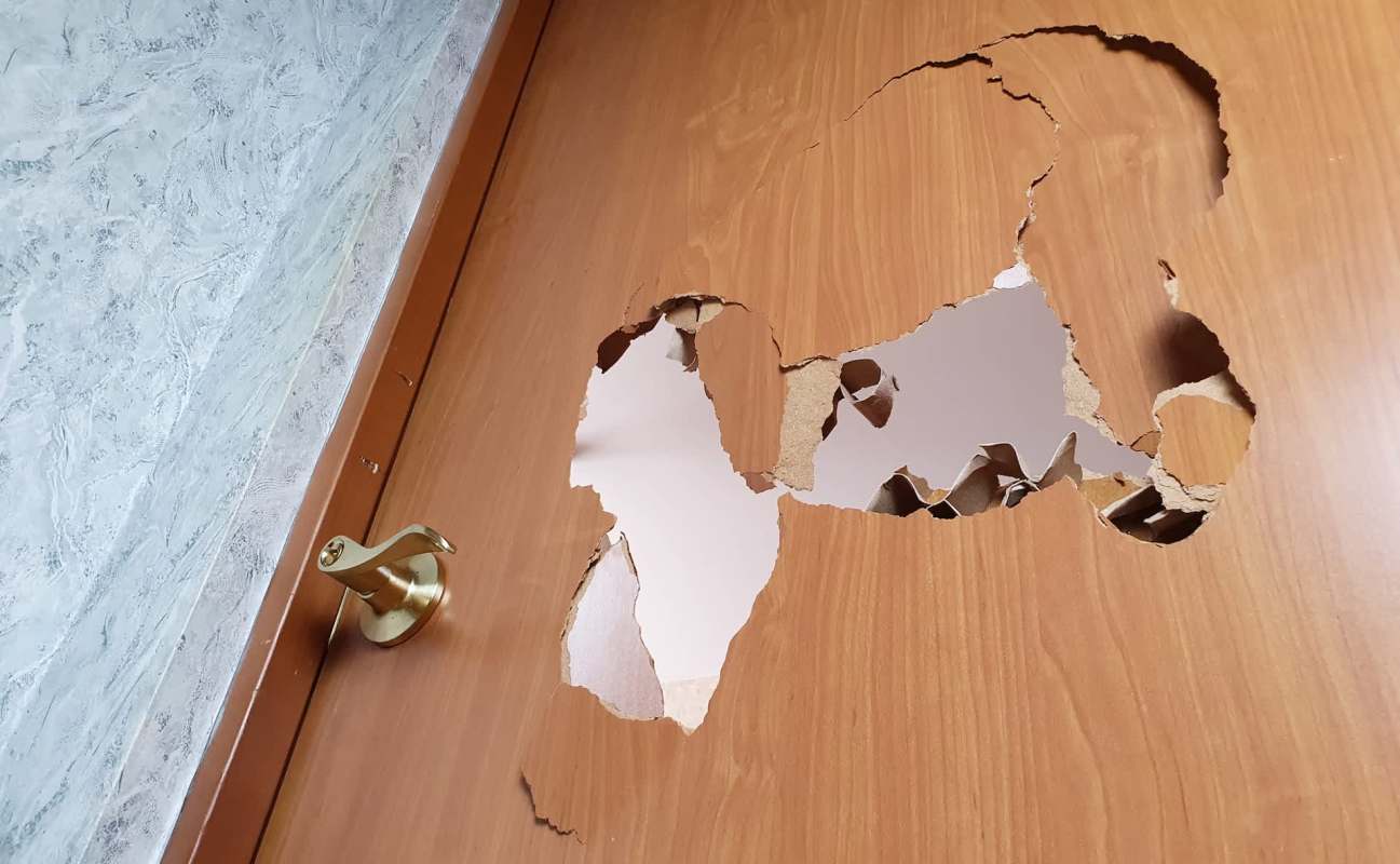 A wooden door with a large, jagged hole near the knob, revealing a light interior.