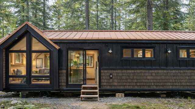 A modern tiny house with a black exterior and copper roof, nestled among tall green trees.