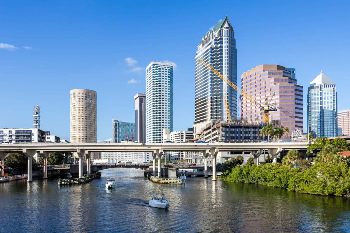 A city skyline with modern skyscrapers alongside a river, featuring boats and a construction site.
