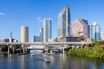 A city skyline with modern skyscrapers alongside a river, featuring boats and a construction site.