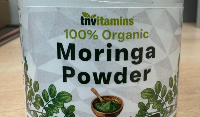 A container of 100% organic moringa powder with a wooden scoop and green leaf illustrations.