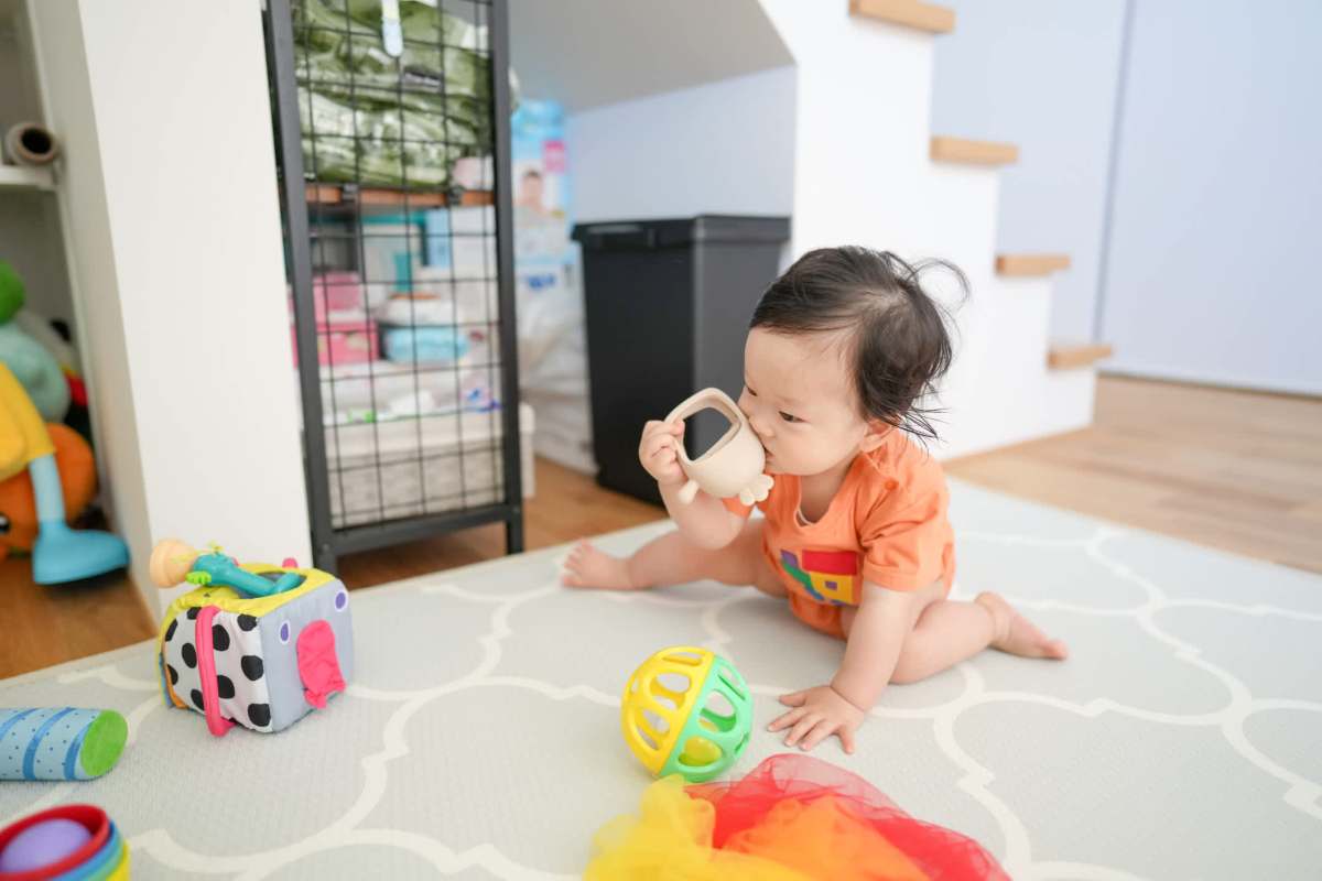A toddler sitting on a playmat, playing with colorful toys and sipping from a small cup.