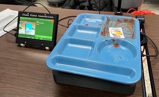 A blue cafeteria tray with remnants of food beside a digital display labeled "Clean Plate Innovations."