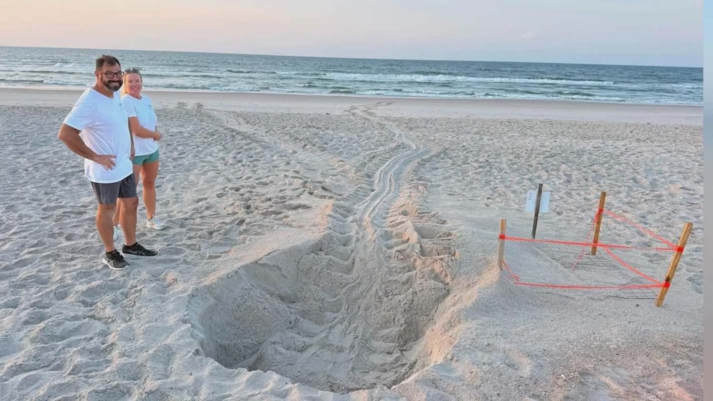 Two people stand on the beach next to a large trench in the sand, with ocean waves visible in the background.