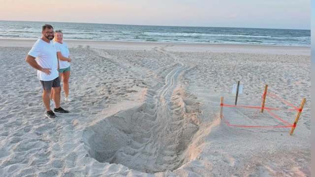 Two people stand on the beach next to a large trench in the sand, with ocean waves visible in the background.