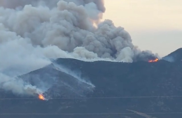 Two wildfires burn on a mountain, producing thick smoke and visible flames against a sky at dusk.