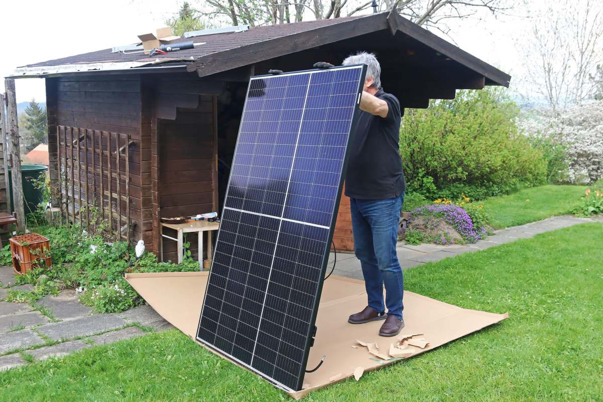 A person is lifting a solar panel next to a wooden shed in a grassy yard.