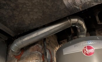 Metal pipes connect to a Rheem water heater in a dimly lit basement with brick walls.