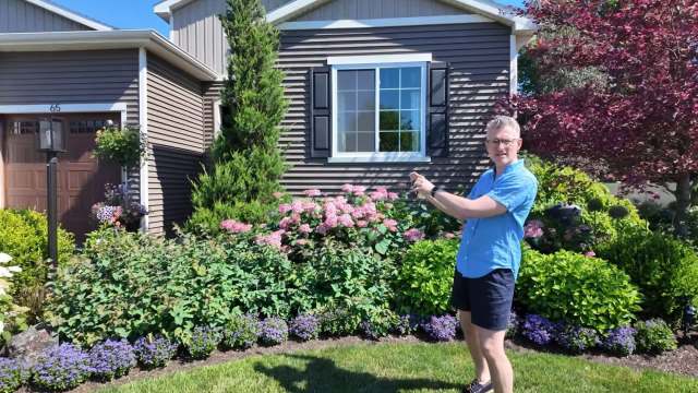 A person stands beside colorful garden flowers in front of a house.