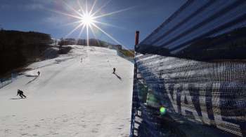 Skiers navigate a snowy slope under a bright sun, with blue safety netting in the foreground.