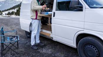 A person cooking at a portable stove next to a van, with a folding chair and scenic mountains in the background.