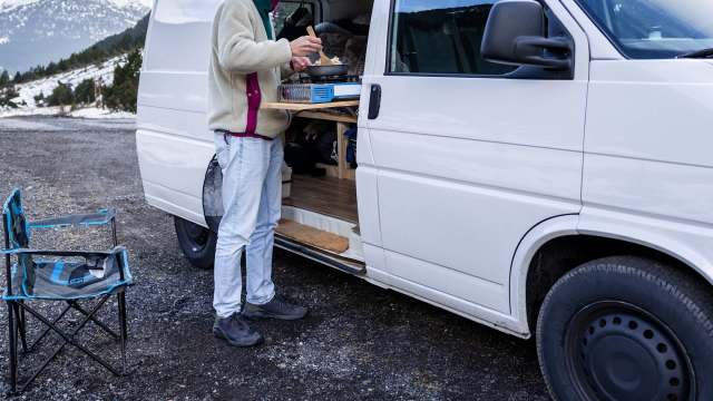 A person cooking at a portable stove next to a van, with a folding chair and scenic mountains in the background.