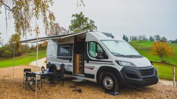 A camper van parked on gravel with a person sitting outside under an awning in a scenic, autumn landscape.